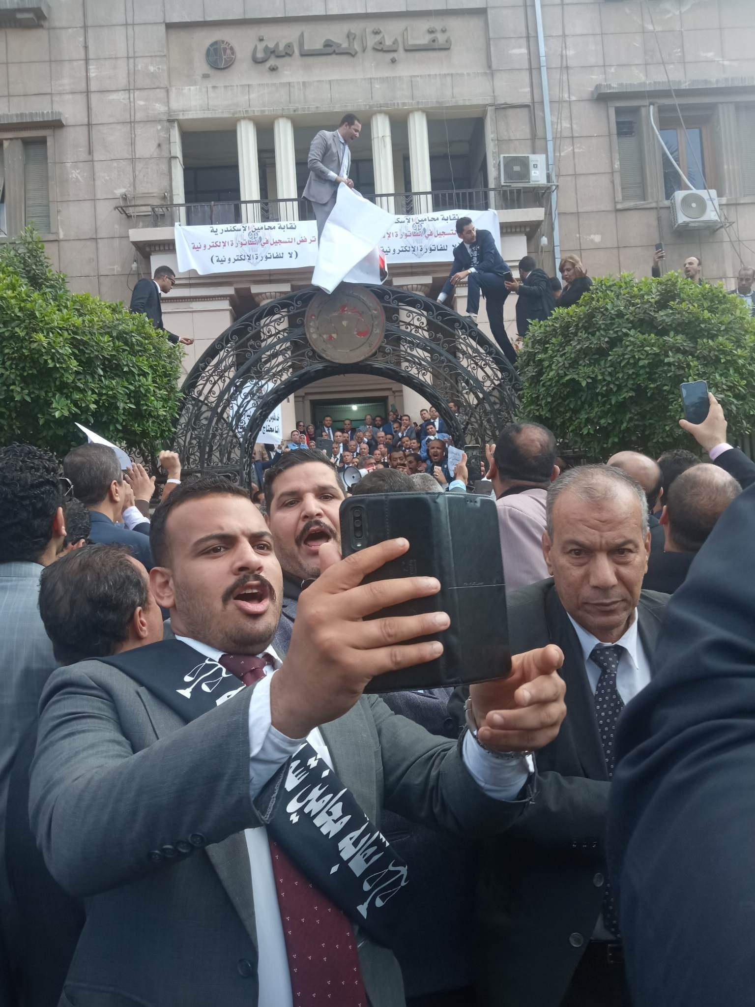 تواصل نقابة محامين شمال القليوبية، مشاركاتها مع القاهرة والمحافظات الاخرى، في وقفات الاحتجاج على إلزامهم بالتسجيل في الفاتورة الإلكترونية، مؤكدة في بيان لها أنها تستشعر الخطر فيما يجرى من محاولة فرض الفاتورة الإلكترونية على المحامين. وأكدت النقابة على رفضها التام لتسجيل المحامين في منظومة الفاتورة الإلكترونية لأن المحاماة مهنة لا تتعلق بتداول السلع أو الاتجار فيها وأن أعمالها هي أعمال قانونية وذهنية. وأصدرت نقابة المحامين بشمال القليوبية بيان لها، عن الاعتراض على منظومة الفاتورة الالكترونية، مؤكدين المشاركة في الوقفة. وجاء في البيان: "نقابة محامين شمال القليوبية:- أولاً - المشاركة في الوقفة السلمية المحددة لها النقابة العامة للقاهرة تضامنا مع باقي النقابات الفرعية وتوفير وسيلة الانتقال الي النقابة العامة من النقابات الفرعية. ثانياً - تقرر وقف التعامل مع جميع خزن محاكم شمال القليوبية يومي 7 و 8/ 2022/12. ثالثاً - الدعوة الي اجتماع جمعية عمومية طارئ السبت المقبل الموافق 10/12/2022, وذلك لمناقشة أخر المستجدات وفقاً لأخر الأوضاع. رابعاً - من يخالف قرار المجلس يحال للتأديب. خامساً - تم إحالة جميع من يخالف القرار الي النقابة العامة. كانت ودعت مصلحة الضرائب مؤخرًا جميع الممولين ومنهم أصحاب المهن الحرة ممن لديهم تعاملات مع المستهلكين النهائيين إلى التسجيل بمنظومة الفاتورة الإلكترونية في موعد أقصاه 15 ديسمبر الجاري. وقال مجدي المهدي نقيب المحامين بشمال القليوبية، المحامون لا يحملون الدولة أية أعباء مالية بل أن المحامين لا تحصل على أي حق من الحقوق كأي مواطن اخر، مؤكدا أنهم لا يحصلون على معاشات من الدولة بل أن دخلهم من عملهم القانوني المباشر فقط. واضاف "المهدي" لن تتردد النقابة في اتخاذ أية إجراءات تكفل منع تطبيق الفاتورة الإلكترونية على المحامين والدفاع عن حقوق أعضاء النقابة. وأكد أحمد رمضان محام وعضو اللجنة النقابية بشمال القليوبية، أن النقابة لا تزال في حالة انعقاد دائم لحين انتهاء الأزمة بالعدول عن قرار تسجيل المحامين بمنظومة الفاتورة الإلكترونية"، مضيفا ان هناك تصعيد في حالة استمرار فشل مفاوضات لجنة المشكلة بين وزارة المالية والنقابة العامة لحل الأزمة. وطالب أحمد رمضان علي بضرورة الغاء قرار تسجيل المحامين بمنظومة الفاتورة الإلكترونية لكون المحاماه رسالة وليست مهنة تجارية. كانت قررت الجمعية العمومية لمحامي مصر اليوم الخامس من ديسمبر ٢٠٢٢م الآتي : ١- الإمتناع التام عن التعامل مع الخزينة بالمحكمة وسداد أية رسوم على مستوى كل محاكم مصر بكل الجزئيات، ويستثنى من ذلك الدعاوى والإجراءات المرتبطة بمواعيد، وكلفت مجالس النقابات الفرعية بتنفيذ ذلك. ٢- حددت موعد الخميس الموافق ٨ ديسمبر ٢٠٢٢م الساعة ١٢ ظهرًا بالنقابة العامة للمحامين لعرض قرار الإضراب عن العمل. ٣- أكدت الجمعية العمومية للمحامين مصر على النقيب العام ومجلسه بضرورة الالتزام بتنفيذ قرارات الجمعية العمومية. ٤- نبهت الجمعية العمومية للمحامين مصلحة الضرائب المصريه بسرعة العدول عن القرار الباطل بإلزام السادة المحامين بالإشتراك بمنظومة الفاتورة الإلكترونية والإيصال الإلكتروني. ٥- نبهت الجمعية العمومية للمحامين على المستشار وزير العدل بأنه لا رسوم إلا بقانون، وأنه عليه وقف قرارات رؤساء بعض المحاكم بفرض رسوم جديدة بناء على تعليمات وبالمخالفة لقانون الرسوم القضائية. ٦- طالبت الجمعية العمومية للمحامين من البرلمان المصري التصدي للمحاولات الباطلة بإخضاع السادة المحامين لمنظومة الفاتورة الإلكترونية والإيصال الإلكتروني. وتوجه شكر للسادة النواب الذين تقدموا بطالبات إحاطة للبرلمان . ٧- طالبت الجمعية العمومية للمحامين من المحكمة الدستورية العليا الفصل في الطعن الدستوري بشأن قانون القيمه المضافة وبطلان إلزام المحامي بضريبة الجدول. وأكدت على الموعد الخميس ٨ ديسمبر ٢٠٢٢م